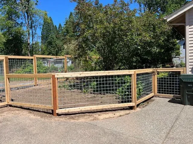 Wooden fenced dog run beside a house, with wire mesh panels and gravel ground.