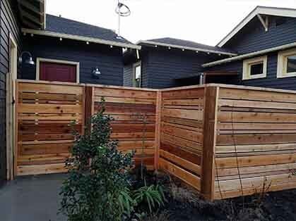 Wooden privacy fence around a small courtyard beside dark blue houses, with a young tree in front.