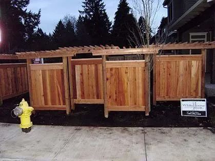 Wooden fenced trash enclosures on a sidewalk beside a yellow fire hydrant at dusk