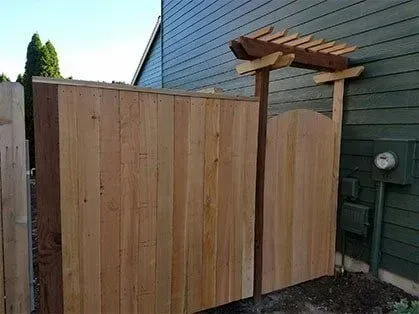 Wooden privacy fence and gate beside a gray house, with a small pergola overhead