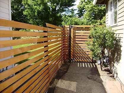 Wooden slat fence lining a narrow concrete side yard beside a house and shrubs.