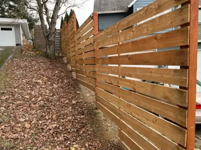 Narrow leaf-covered yard beside a wooden fence and house with a tree in the background