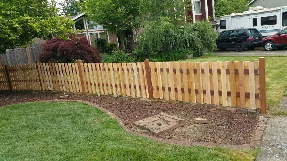 Wooden fence along a lawn and garden bed in a suburban yard, with houses and parked cars in the background