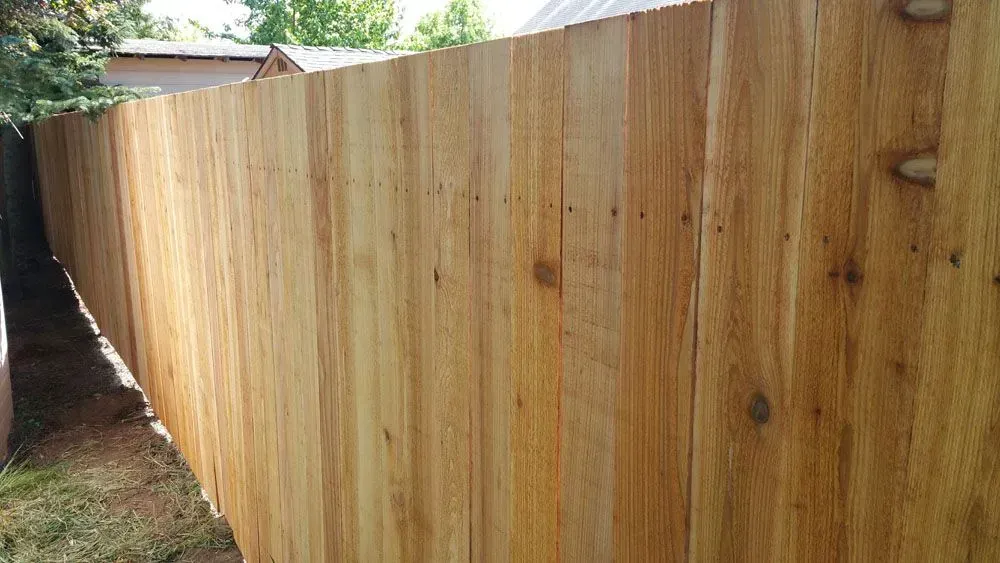 Wooden privacy fence along a narrow side yard, with trees and a house roof visible above.