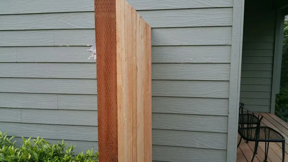 Wooden planter or privacy screen beside a gray house wall with shrubs at the base.