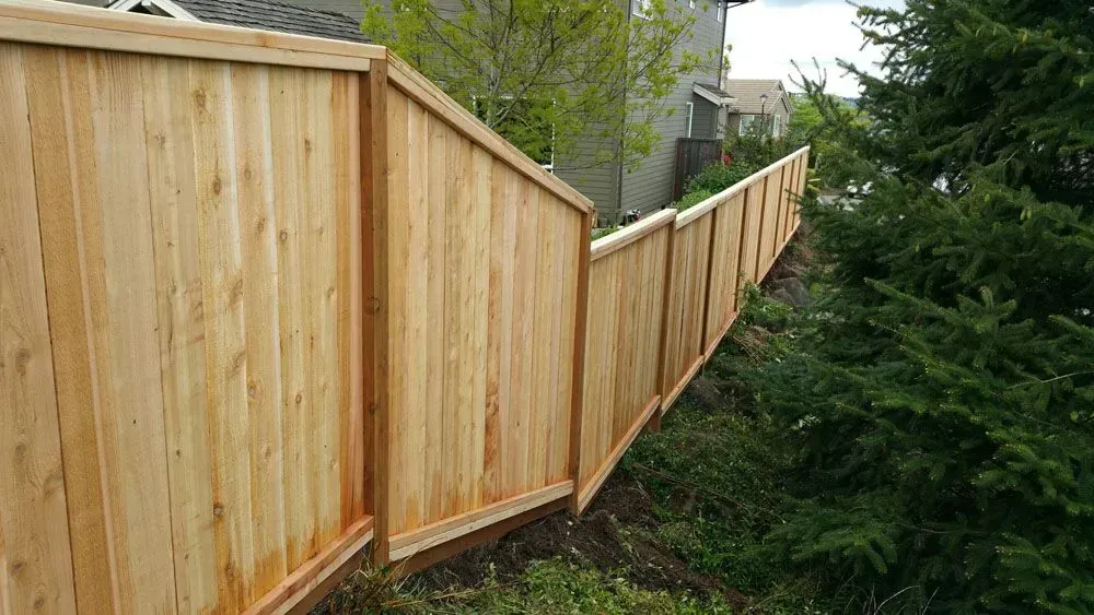 Tall wooden privacy fence along a sloped backyard beside trees and neighboring houses.