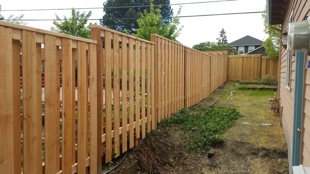 Wooden privacy fence along a narrow backyard path beside a house, with grass and dirt on the ground.