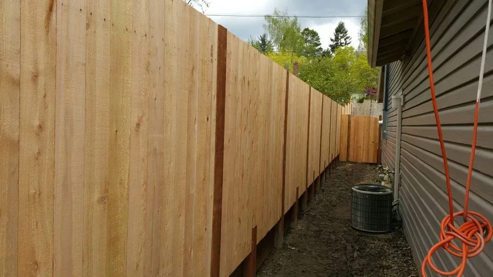 Tall wooden fence along a narrow gravel walkway beside a house, with orange extension cords on the right.