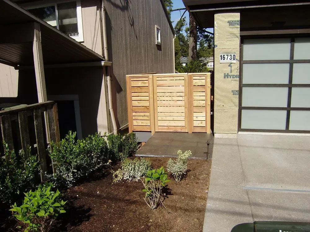 Residential entrance with wooden gate, concrete path, and small landscaped garden beside a building