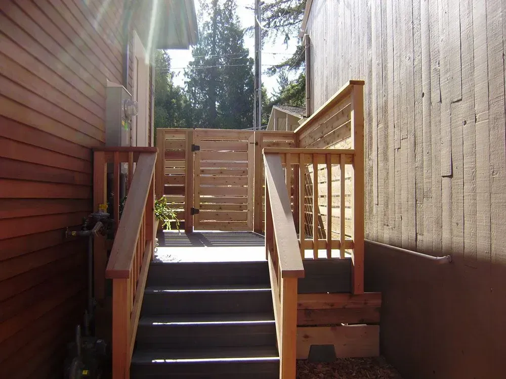 Wooden outdoor stairs between two buildings leading to a fenced landing in bright sunlight