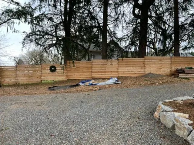 Gravel yard with a wooden fence, bare trees, and a stone border under an overcast sky