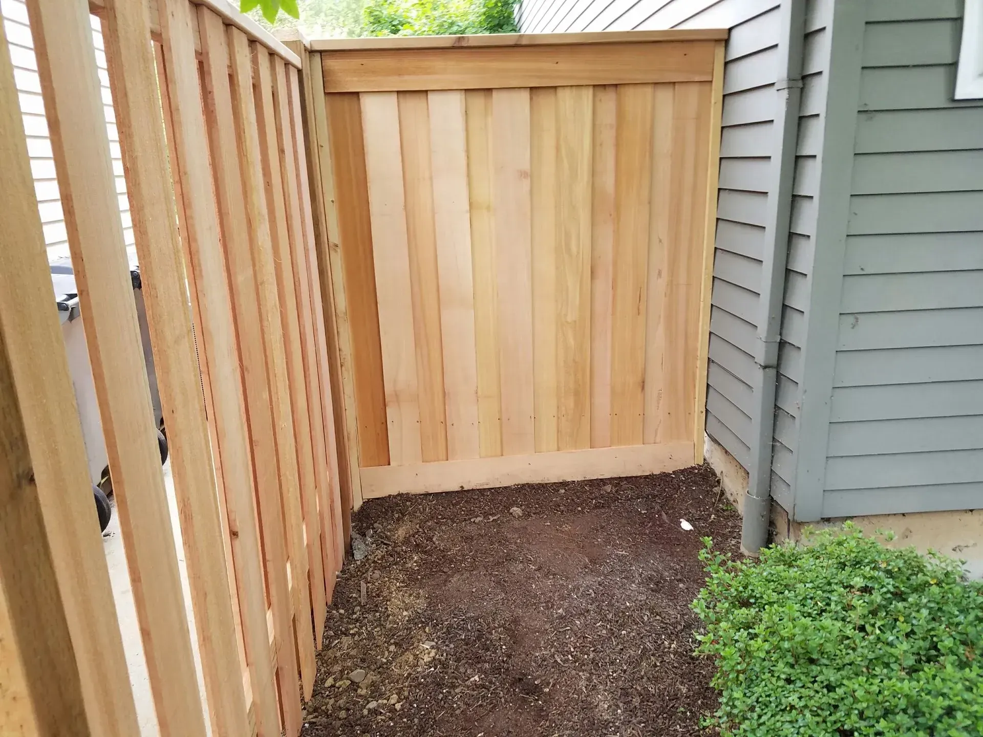 Small fenced backyard corner with a wooden gate, dirt ground, and a bush beside a house.