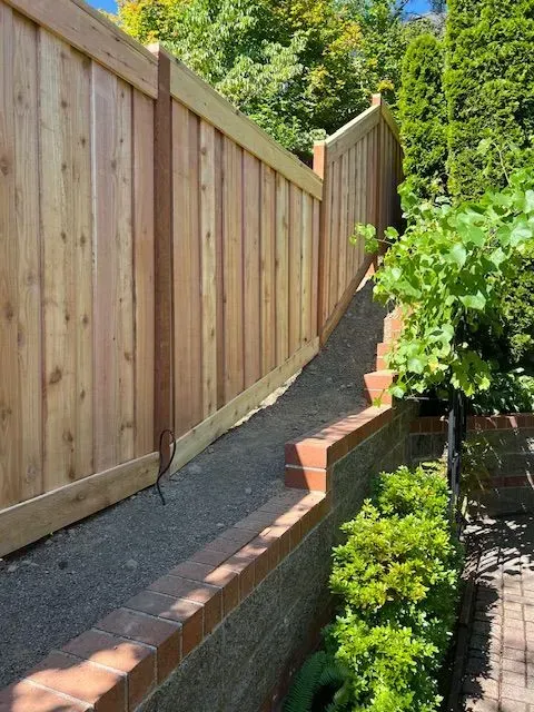Tall wooden fence along a narrow brick-and-concrete garden path beside green shrubs and trees