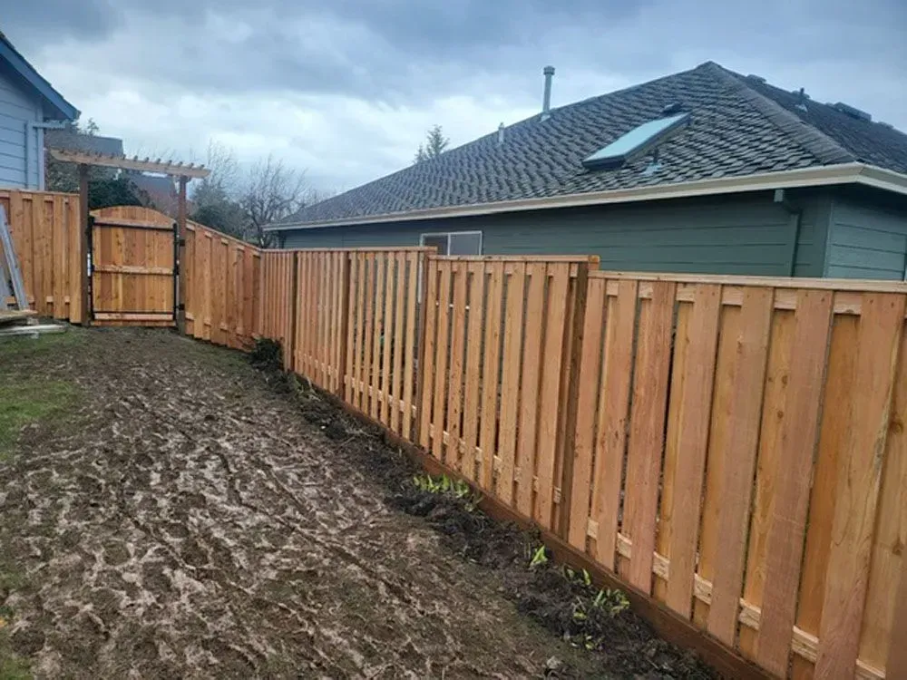 Wooden fence along a muddy backyard beside a gray house under an overcast sky