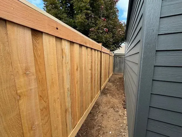 Narrow dirt path between a wooden fence and gray house siding leading to a backyard gate.