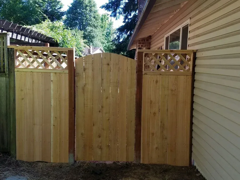 Wooden backyard fence and gate beside a beige house, with trees in the background