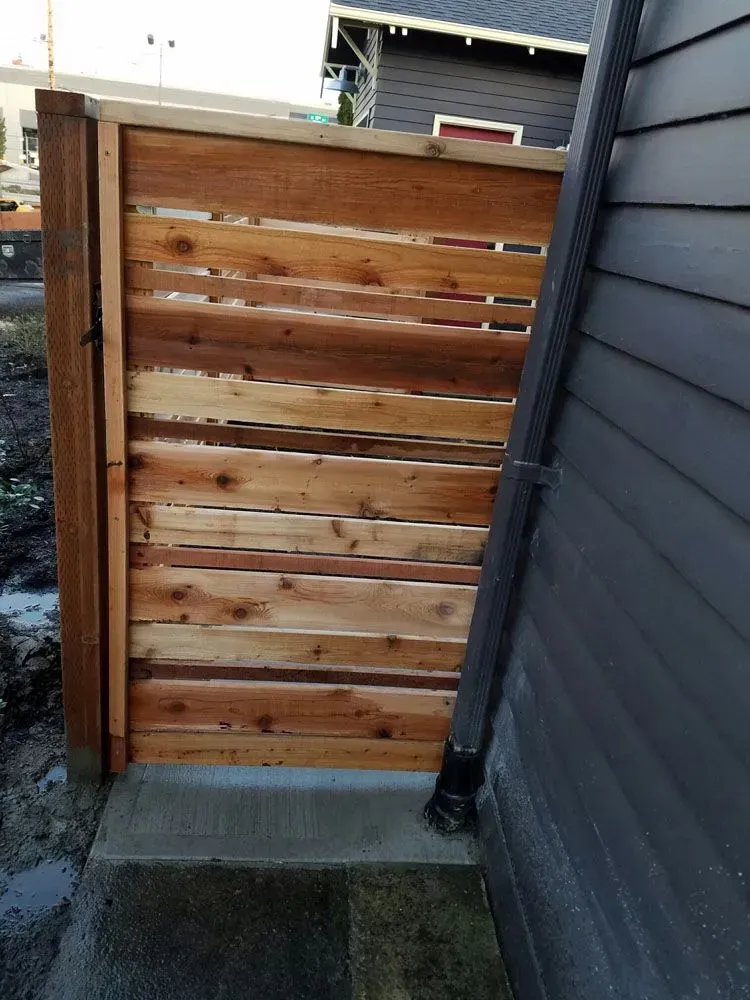 Narrow wooden gate with horizontal slats between a gray siding wall and a fence over a concrete path