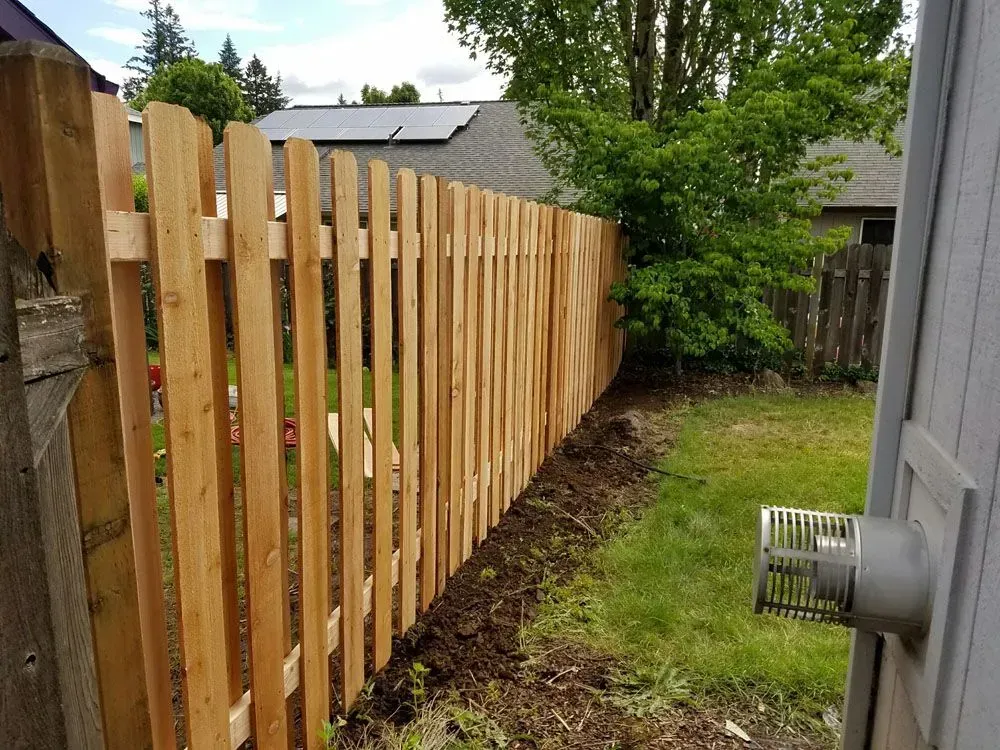 Wooden fence along a narrow grassy side yard beside a gray house and trees