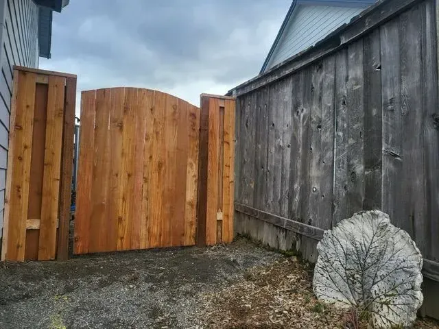 Wooden gate and fence beside a weathered gray wall, with a white rock in the gravel foreground
