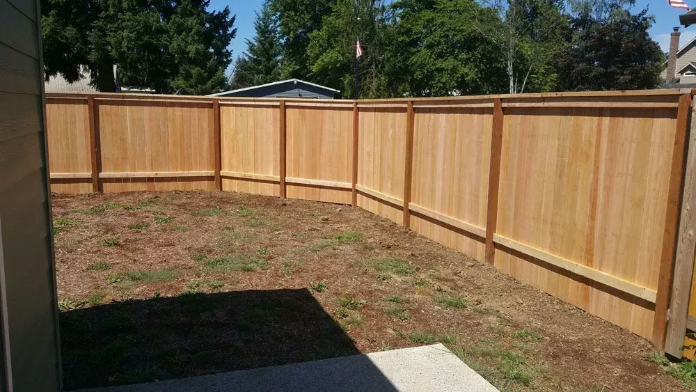 Corner of a fenced backyard with a wooden fence, patchy grass, and a concrete patio in the foreground