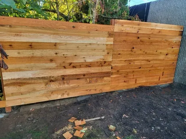 Wooden privacy fence with mixed light and dark planks, partially built along a gravel yard.