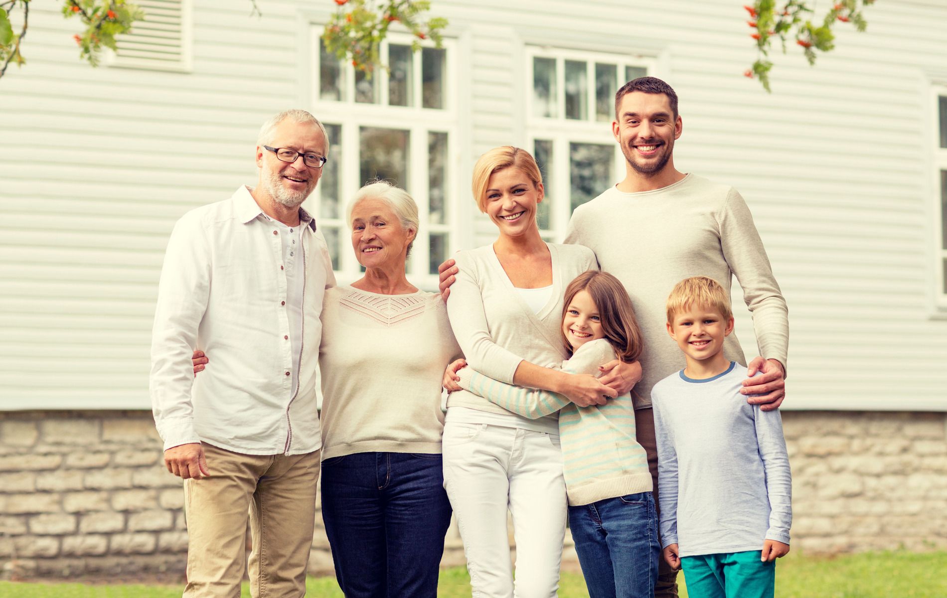 Family portrait in front of a white house; grandparents, parents, and two children smiling.