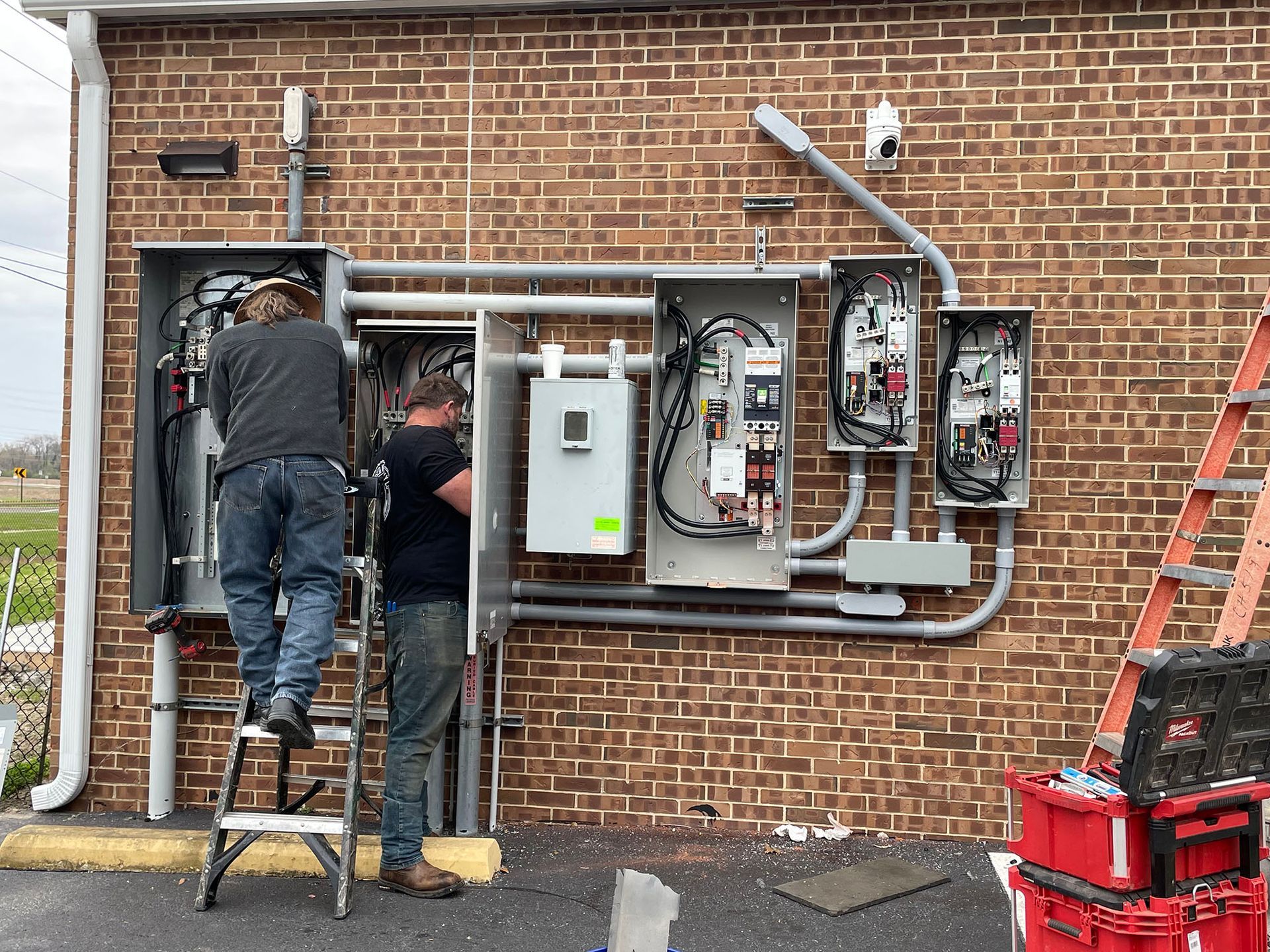 Two men are working on an electrical box on the side of a brick building.
