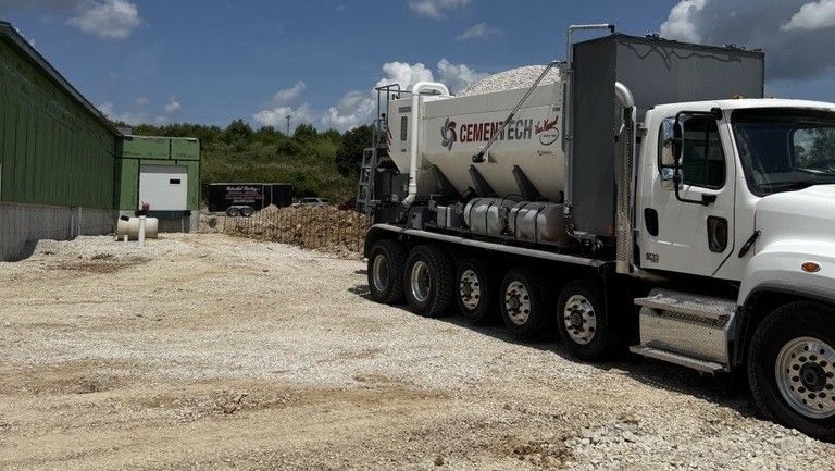 A white cement truck parked beside a building, delivering a load. The ground is covered in gravel, with a dog visible by the building entrance.