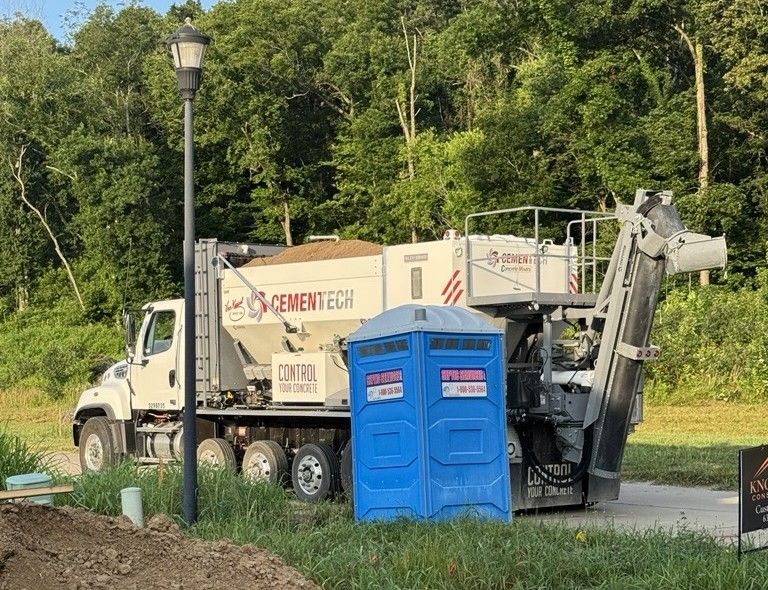 A white cement truck and a blue portable toilet are parked on a road near green trees. A black street lamp is in the foreground.