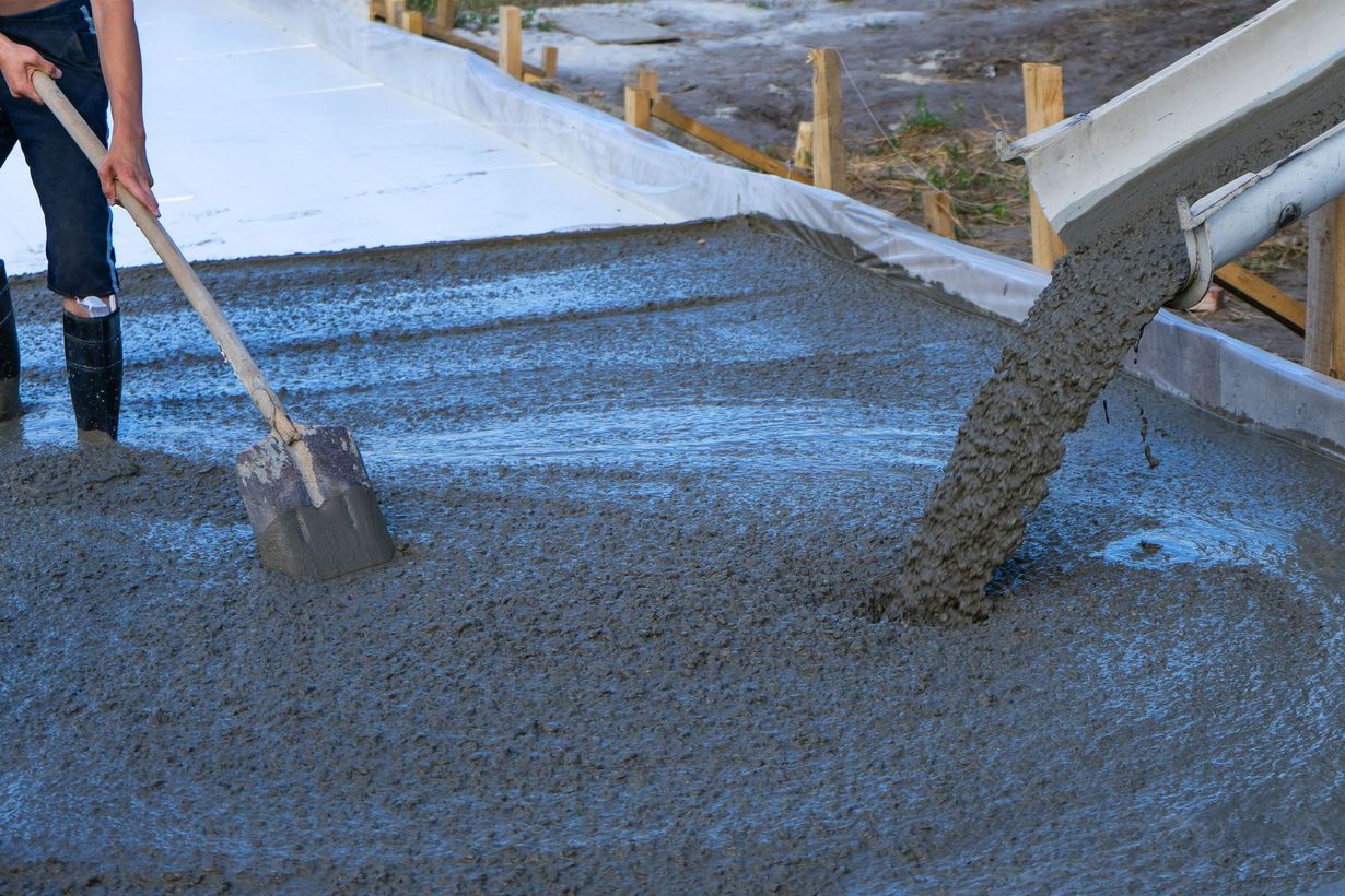 A worker pours concrete from a chute into a form while another spreads it with a shovel on a construction site.