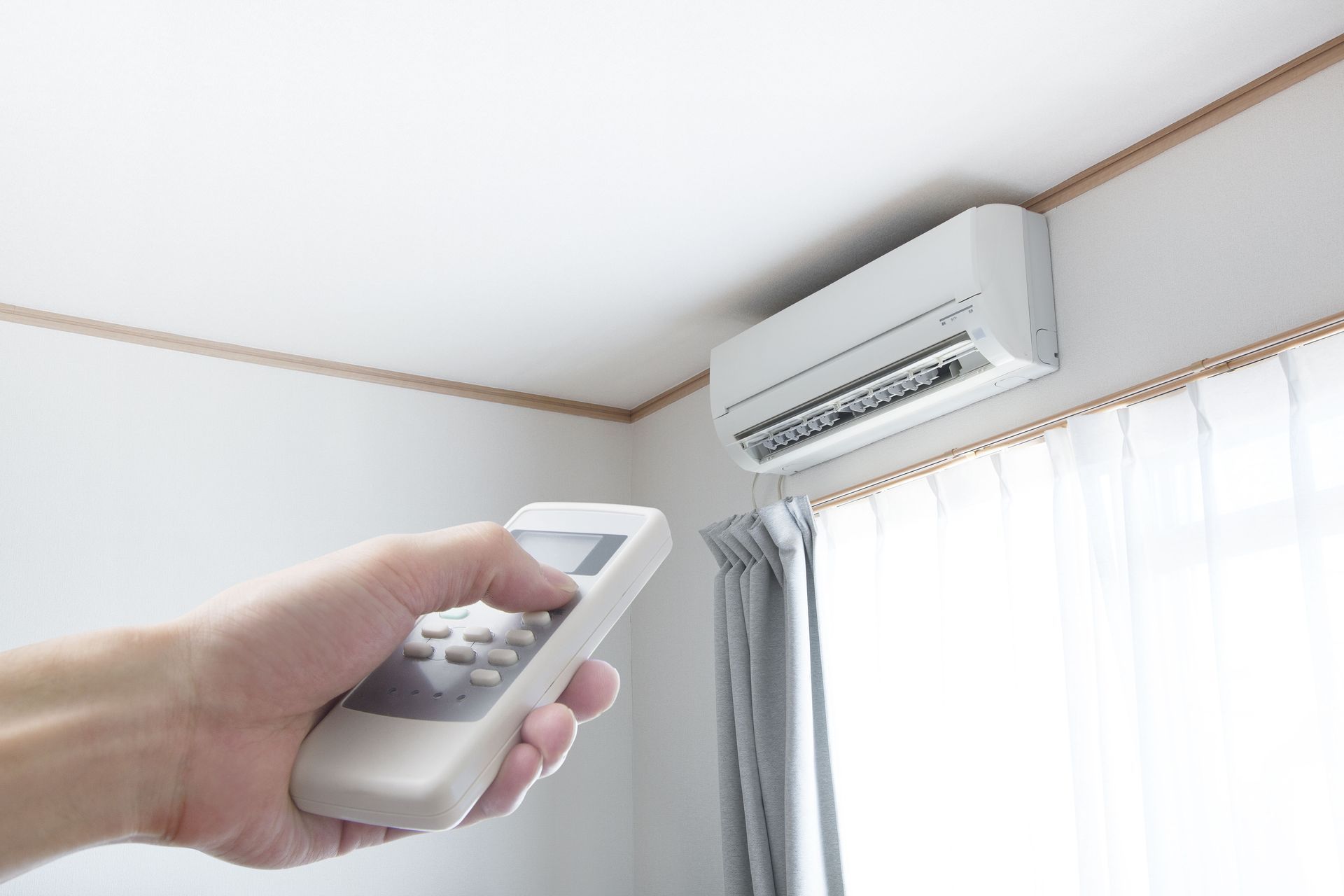 Hand holding AC remote, pointing at wall-mounted AC unit near a window with curtains.