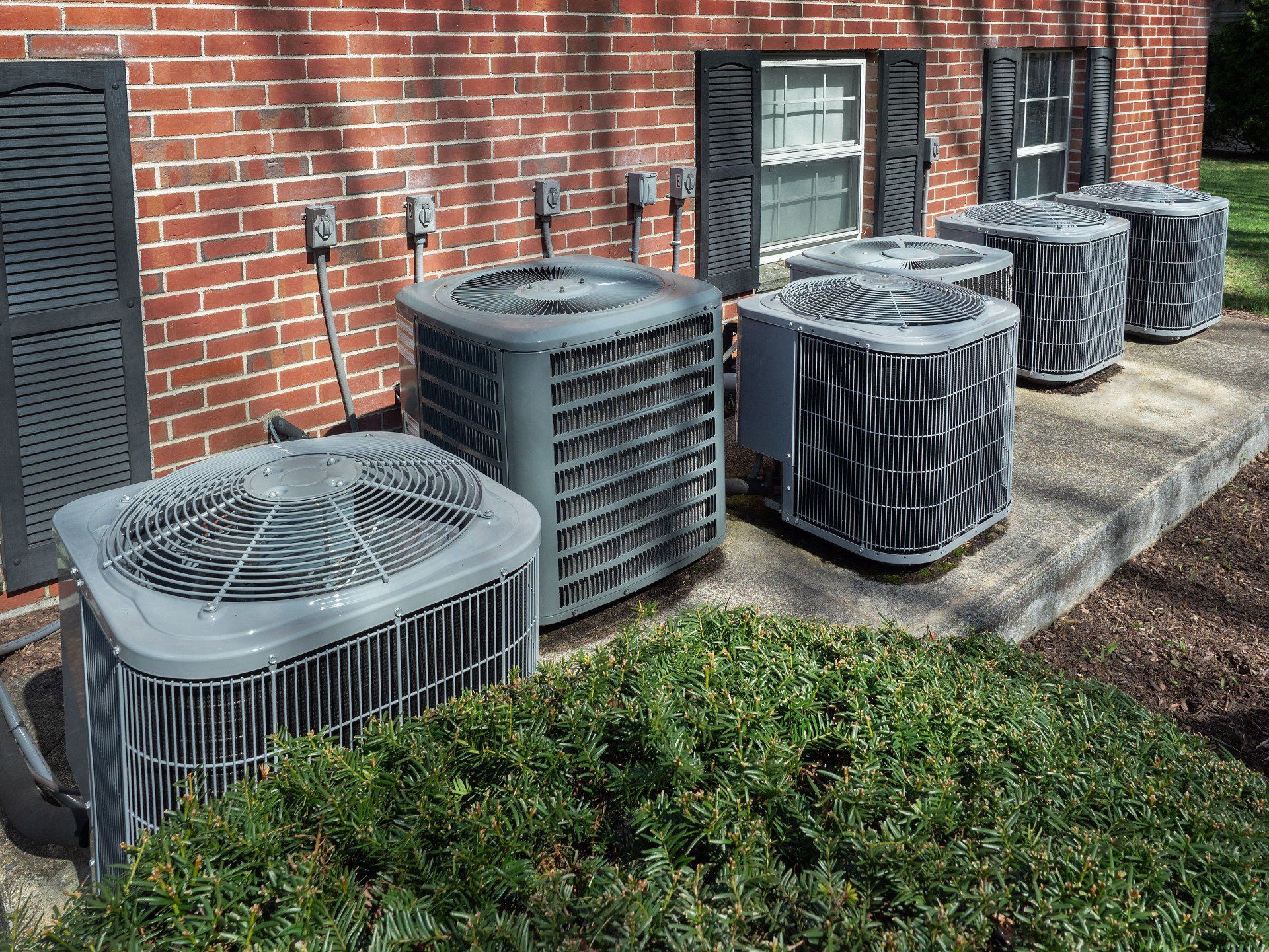 Several air conditioning units lined up against a brick building with green bushes in front.