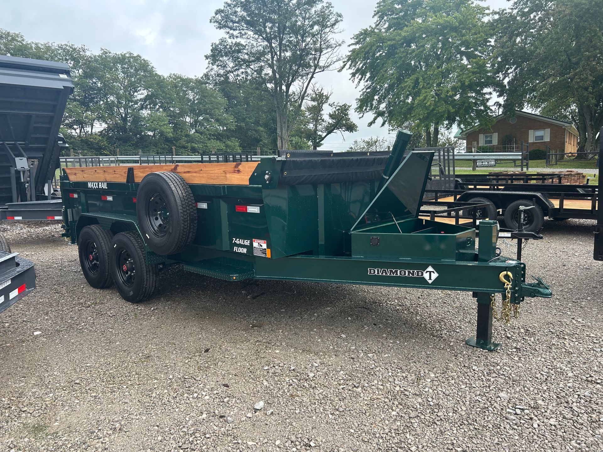 Green dump trailer with raised rear gate parked on gravel lot outdoors