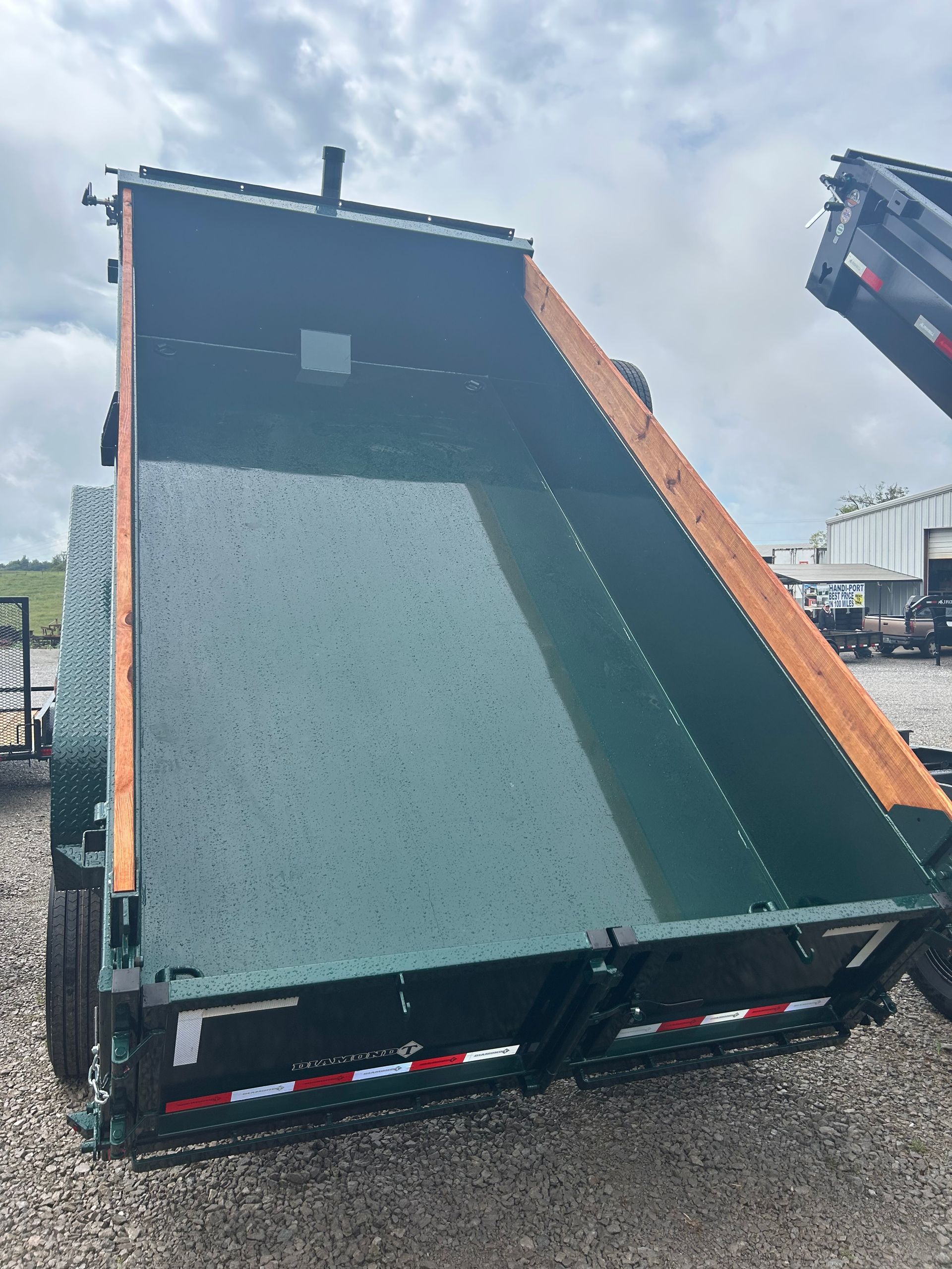 Large green dump trailer bed tipped upright on a gravel lot, with a raised truck arm visible at right.