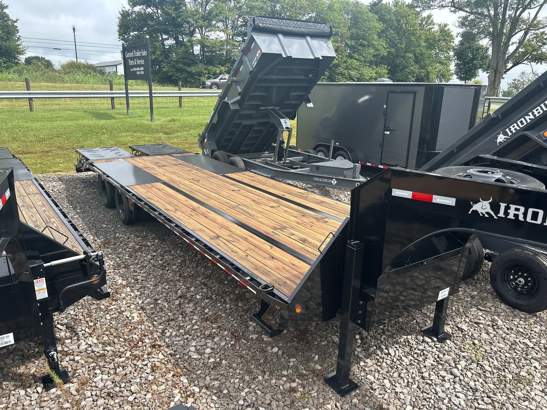 Black dump trailer tipped on gravel, with a raised rear bed and wood plank side panel near a road.