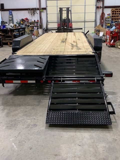 Black flatbed trailer with a wood deck and extending ramp in a workshop.