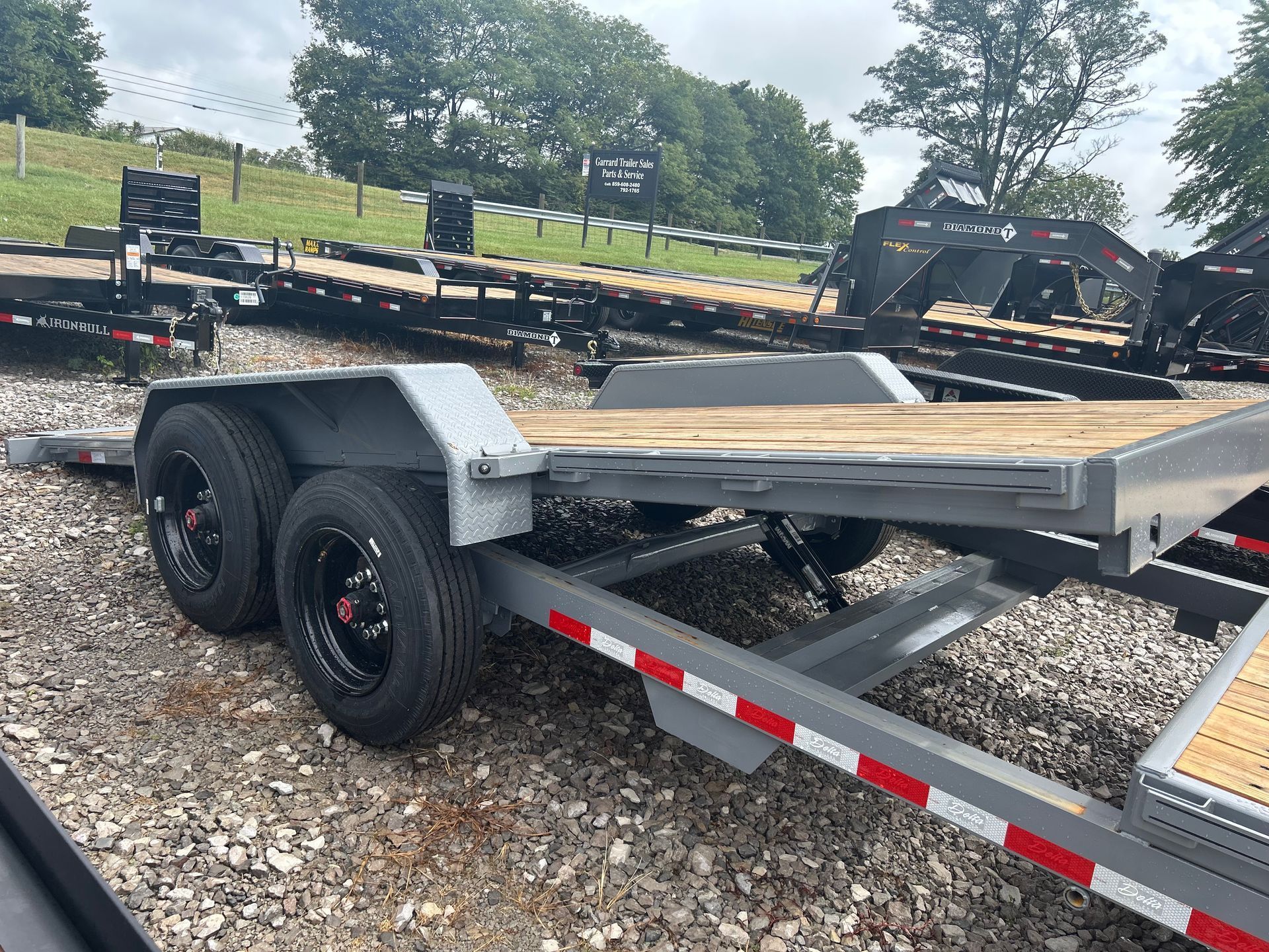 Gray dual-axle flatbed trailer with black wheels parked on gravel in a lot, with trees and other trailers behind.