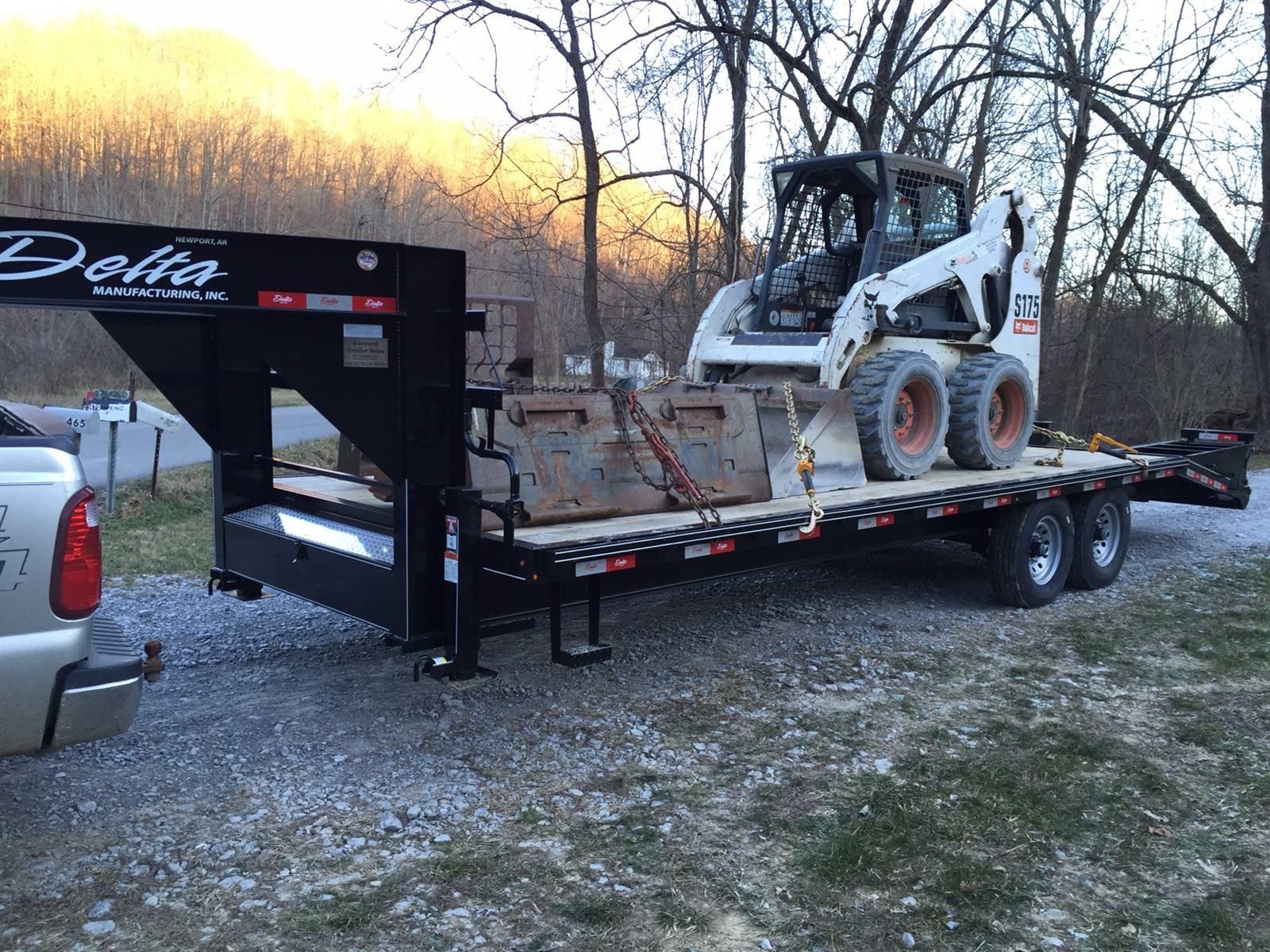 Black trailer loaded with a Bobcat skid-steer and concrete blocks parked on gravel, with trees in the background.