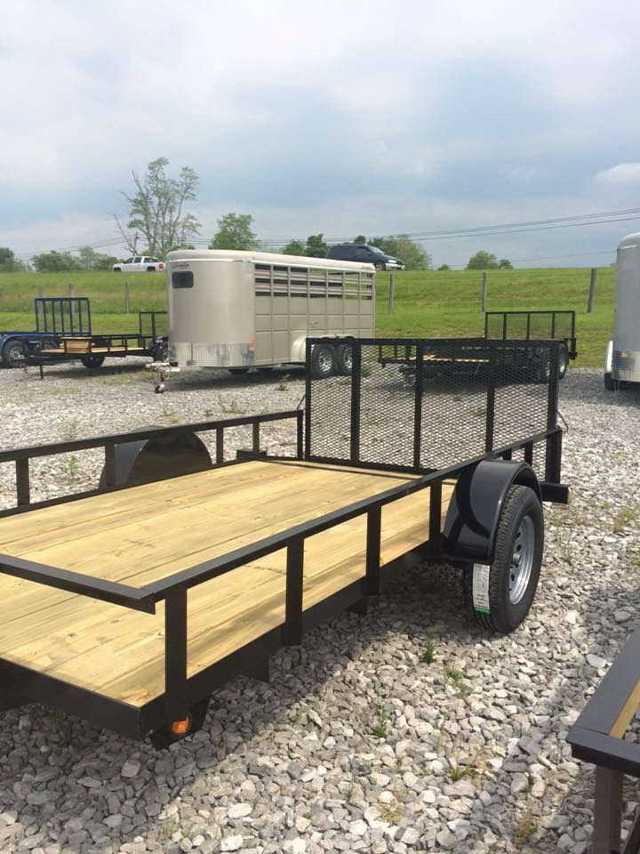 Black and tan utility trailer with mesh railing, parked on gravel in a field with other trailers.