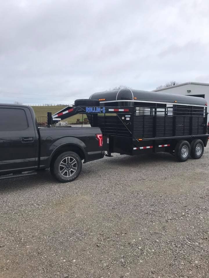 Black pickup truck towing a black livestock trailer on a gravel surface under a cloudy sky.