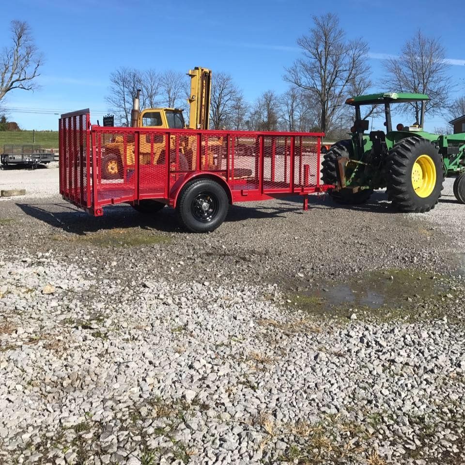 Red trailer hitched to a green tractor on a gravel lot; a yellow piece of heavy machinery in the background.