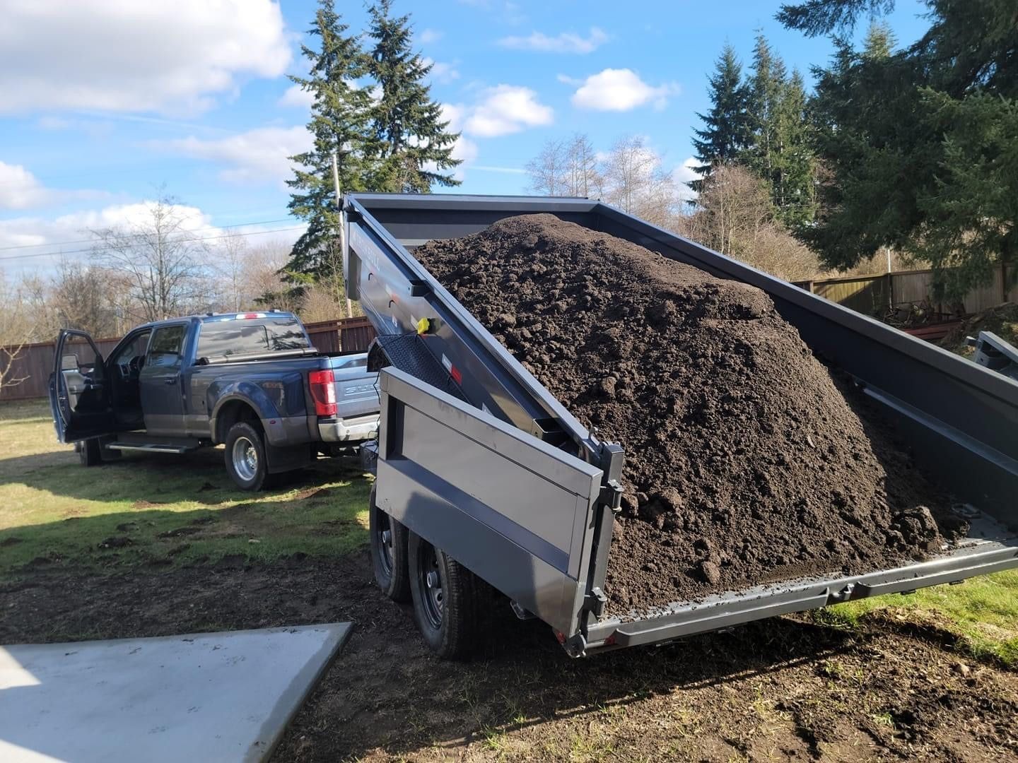 Dark truck bed filled with soil being unloaded. Gray trailer, blue truck, and green grass are visible.