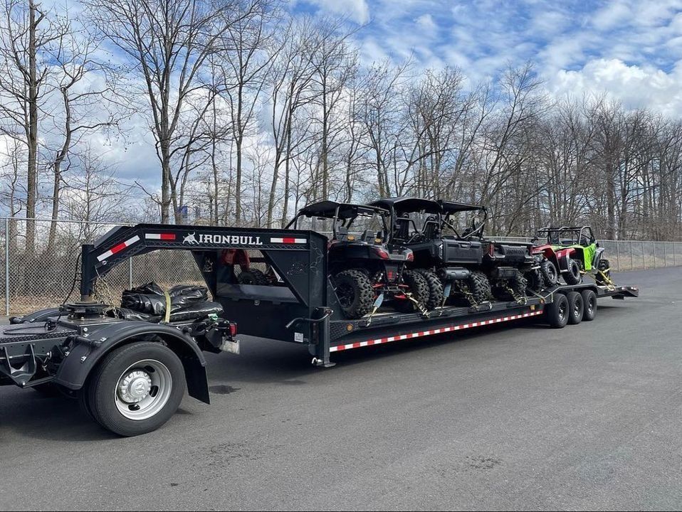 Black trailer hauling several ATVs on a paved road, against a backdrop of trees and a cloudy sky.