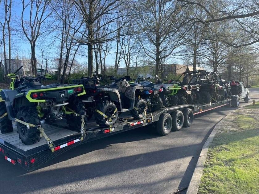 ATVs loaded on a trailer. Various colors, parked on a road, trees in the background.