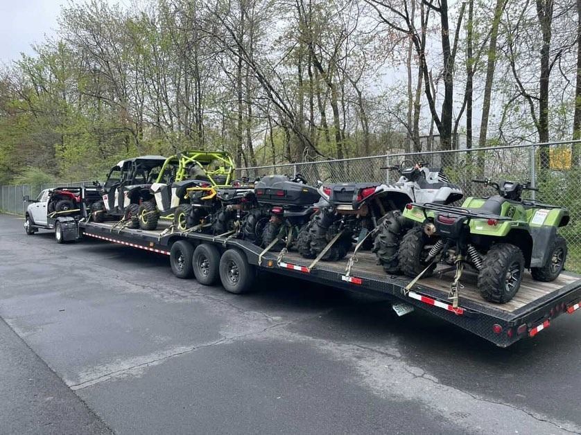 A flatbed trailer loaded with ATVs and a side-by-side vehicle. It's parked on asphalt next to a chain-link fence.