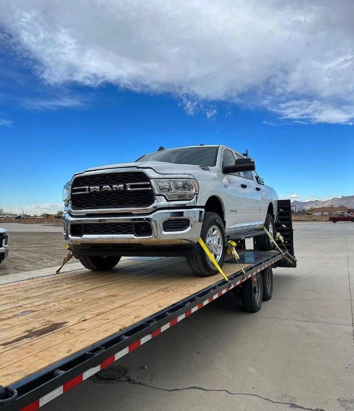 White Ram pickup truck secured on a flatbed trailer, under a partly cloudy blue sky.