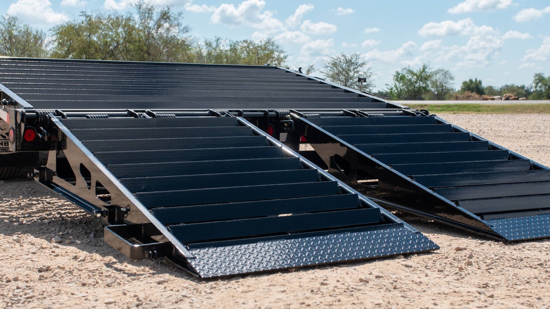 Black ramps extended from a trailer on a gravel surface outdoors.