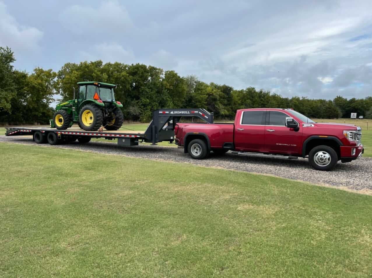 Red pickup truck towing a flatbed trailer with a green John Deere tractor on a grassy area.