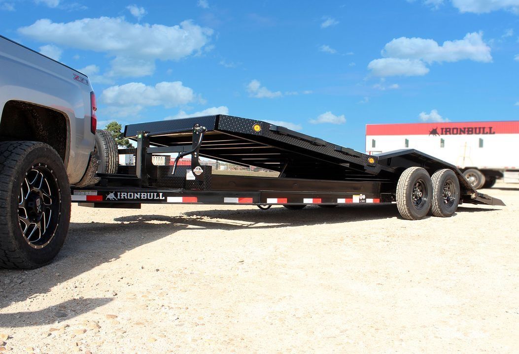 Truck towing a black trailer with a ramp, parked on gravel against a blue sky.