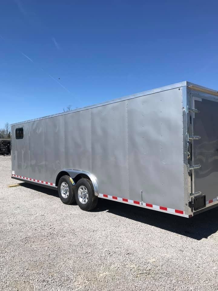 Silver enclosed trailer with two axles parked on gravel under a blue sky.